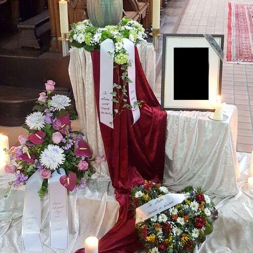 Gedenkaufstellung mit Blumen, Kerzen und einem leeren Rahmen auf einem silbernen Tuch in einer Kirche.
