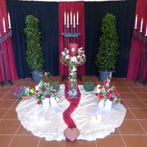 Dekorativer Altar mit roten und schwarzen Stoffen, Kerzen, Blumenarrangements und einer großen roten Kerze in der Mitte.