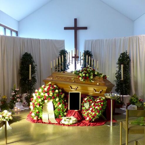 Raum mit einem goldenen Sarg, umgeben von Blumen und Kerzen, im Hintergrund ein Kreuz und Vorhänge.