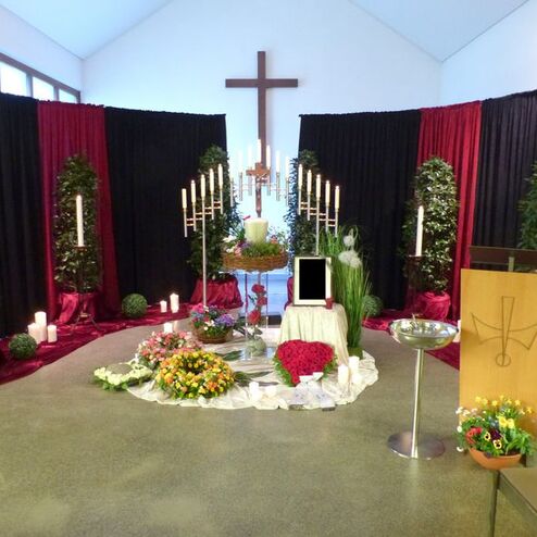 Dekorierte Trauerhalle mit Kre cross, Kerzen, Pflanzen und Blumenarrangements auf einem Tisch.
