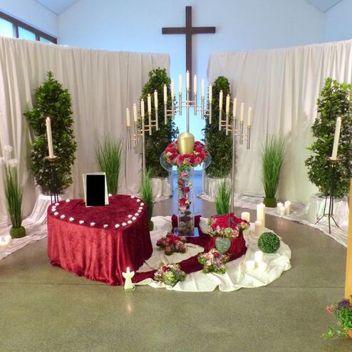 Dekorierte Gedenkstätte mit Blumen, Kerzen und einem Kreuz vor einem hellen Hintergrund in einer feierlichen Umgebung.