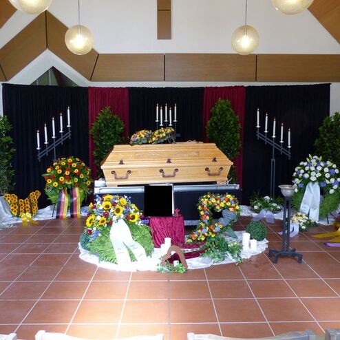 Trauerfeier mit einem offenen Holzsarg, umgeben von Blumenarrangements und Kerzen in einem würdevollen Raum.