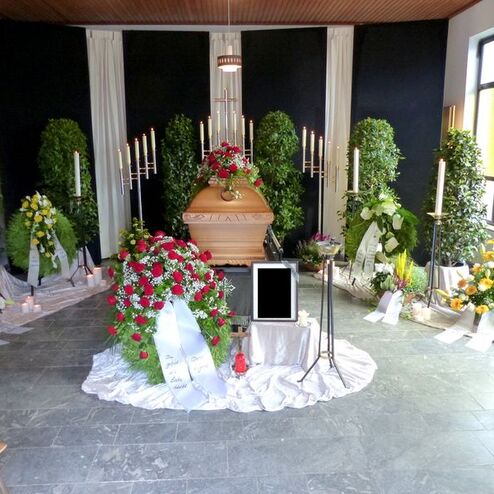 Dekorierter Raum mit einem Sarg, umgeben von Blumen, Kerzen und Pflanzen für eine Trauerfeier.