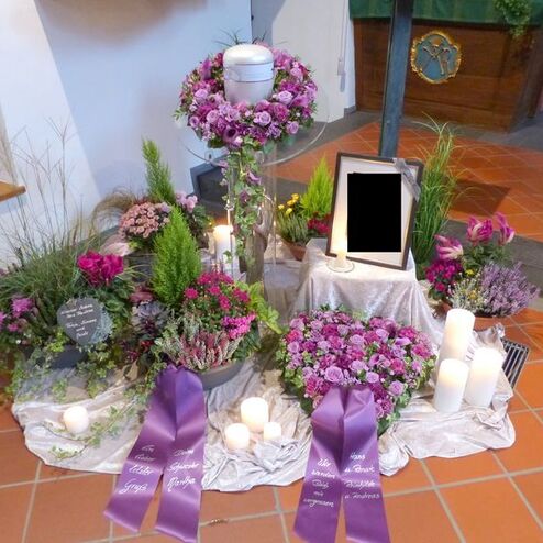 Gedenkstätte mit Blumenarrangements, Kerzen, einer Urne und einer leeren Bilderrahmen auf einem silbernen Tuch.