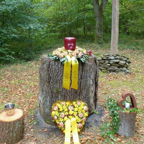 Gedenkstätte im Wald mit einem Baumstumpf, Kerze, Blumenkranz und Steinhaufen, umgeben von herbstlichem Laub.