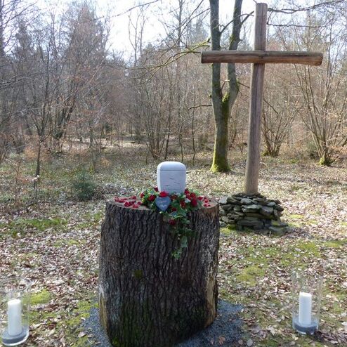 Gedenkstätte in einem Wald, bestehend aus einem Baumstumpf mit Grabstein, Holz-Kreuz und Blumenarrangement.