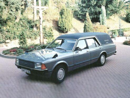 Silberner Leichenwagen parkt auf einem gepflasterten Weg, umgeben von Bäumen und Sträuchern.