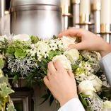 Hände binden frische Blumen und grüne Blätter zu einem Kranz um eine silberne Kerze auf einem Tisch.