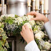 Hände binden frische Blumen und grüne Blätter zu einem Kranz um eine silberne Kerze auf einem Tisch.