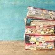 Stapel von Aktenordnern mit farbigen Haftnotizen vor einem hellblauen Hintergrund auf einem Holztisch.