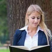 Junge Frau mit langen blonden Haaren liest in einem Buch vor einem großen Baum im Freien.