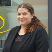 Frau mit_lockigem Haar in schwarzer Bluse und Blazer steht vor einer Glasfassade mit gelbem Logo.