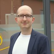 Ein lächelnder Mann mit Brille trägt einen Blazer und ein T-Shirt, steht vor einer Fensterfront.