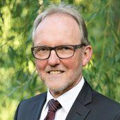 Portrait eines Mannes mit Brille und Anzug vor grüner, unscharfer Pflanzenkulisse.