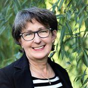 Porträt einer lächelnden Frau mit Brille, grau melierten Haaren, vor grünen Weidenzweigen. Sie trägt ein schwarzes Jackett.