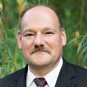 Porträt eines Mannes mit Schnurrbart in Anzug vor grüner, natürlicher Kulisse.